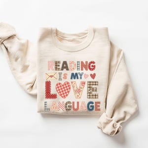 Może przedstawiać: Kremowa bluza z napisem "READING IS MY LOVE LANGUAGE" w stylu patchwork. Tekst zawiera różne wzory, w tym kratę, kwiaty i grochy, z akcentami serca i koperty.