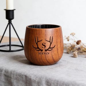 May include: A wooden tumbler with a dark interior and a natural wood grain exterior. The tumbler features a deer antler design with the letter "S" in the center and the name "STEVEN" below. The tumbler is sitting on a gray surface.