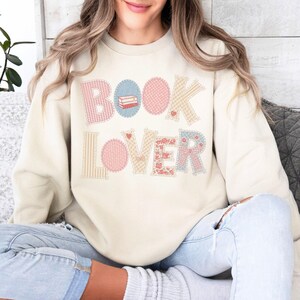 May include: Cream-colored sweatshirt with the words "BOOK LOVER" in patchwork letters. The letters are in various patterns and colors, including pink gingham, floral, and striped designs. A stack of books is in the letter "O".