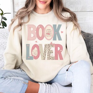 May include: Cream-colored sweatshirt with the words "BOOK LOVER" in a mix of pink, blue, and patterned letters. The "O" in "BOOK" features a stack of books. Small hearts are scattered around the text.