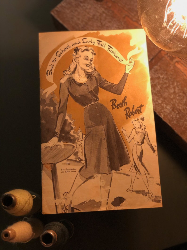 May include: A vintage advertisement for Berth Robert featuring a woman in a gray dress and a white blouse. The text reads "Back to School and Early Fall Fashions".