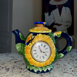 May include: A decorative ceramic teapot with a clock face. The teapot is painted with a sunflower design in yellow, green, and blue. The clock face has black Roman numerals and red hands. The handle and spout are dark blue.