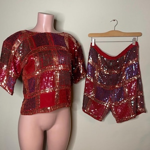 May include: A two-piece sequined outfit in red, purple, and gold. The top has short sleeves and a square pattern. The matching shorts are on a hanger. The outfit is displayed on a mannequin.