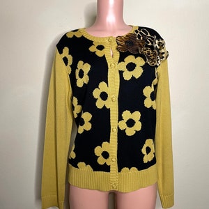 May include: A yellow cardigan with a black floral pattern and a leopard print brooch. The cardigan has a button-up front and long sleeves.
