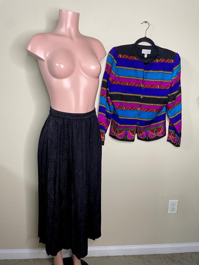 May include: A black paisley print skirt and a colorful button-up jacket with a black collar and gold trim are displayed on a mannequin. The jacket features horizontal stripes in blue, purple, and red.