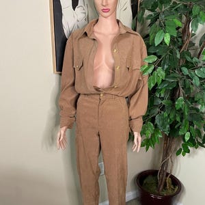 May include: A tan two-piece set consisting of a long-sleeved button-down shirt and matching trousers. The shirt features a collar, two chest pockets, and gold-toned buttons. Both the shirt and trousers are made of a textured fabric with subtle patterns.