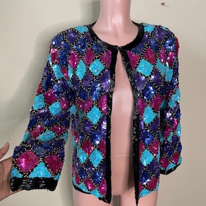 May include: A black sequined jacket with a diamond pattern in shades of turquoise, purple, and pink. The jacket has a front closure and long sleeves.