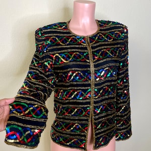 May include: Black jacket with gold trim and colorful sequin embellishments. The jacket features horizontal gold stripes and interwoven sequin patterns in red, green, blue, and gold. The jacket is displayed on a mannequin.