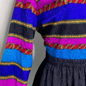 May include: A vintage blouse and skirt set. The blouse features horizontal stripes in blue, purple, black, and red with gold rope-like detailing. The skirt is black with a paisley pattern. The outfit is made of a silky material.