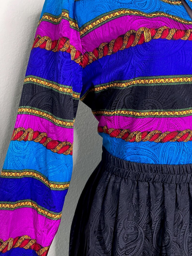 May include: A vintage blouse and skirt set. The blouse features horizontal stripes in blue, purple, black, and red with gold rope-like detailing. The skirt is black with a paisley pattern. The outfit is made of a silky material.