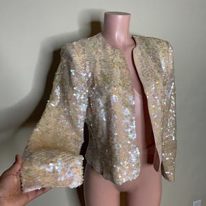 May include: A cream-colored sequined jacket with long sleeves and a V-neck. The sequins are iridescent and create a shimmering effect.