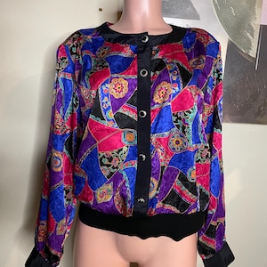 May include: A vibrant, vintage bomber jacket with a colorful patchwork design. The jacket features a black collar, button-up front, and cuffs. The patchwork includes pink, blue, purple, and black geometric shapes with floral accents. The bottom hem is black.