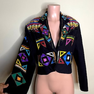 May include: Black cropped blazer with colorful geometric beadwork on the shoulders, lapels, and sleeves. The blazer features a V-neck and long sleeves. The beadwork includes red, yellow, green, blue, and purple accents.