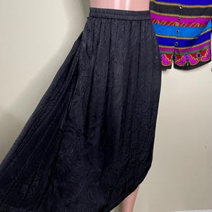 May include: A black, knee-length skirt with an elastic waistband and a paisley pattern. The skirt is made of a silky material and has a flowing, asymmetrical hemline. A colorful patterned top is partially visible in the background.