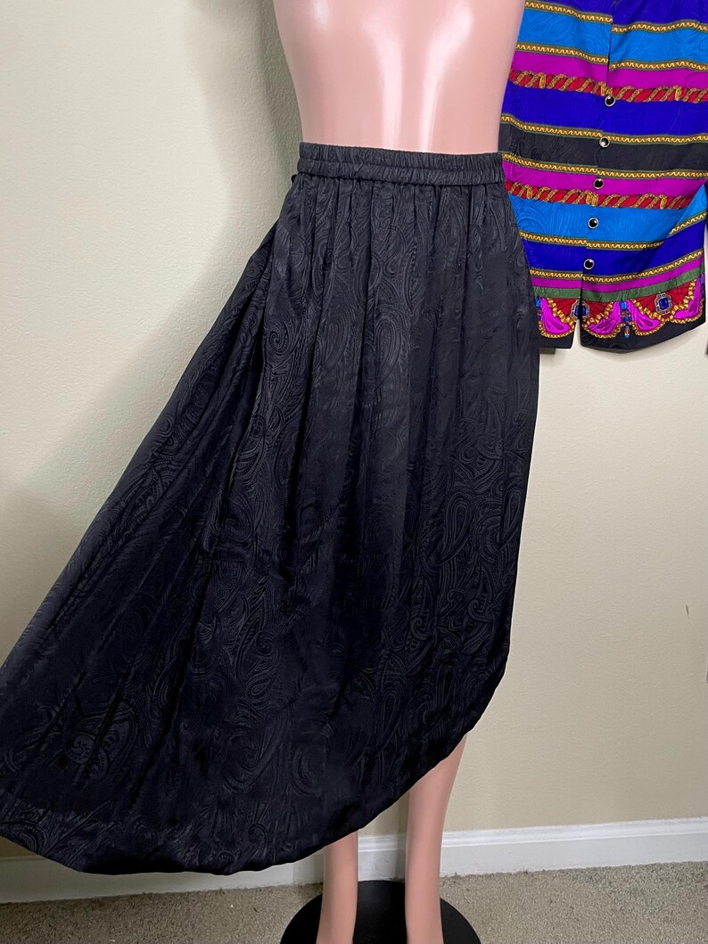 May include: A black, knee-length skirt with an elastic waistband and a paisley pattern. The skirt is made of a silky material and has a flowing, asymmetrical hemline. A colorful patterned top is partially visible in the background.