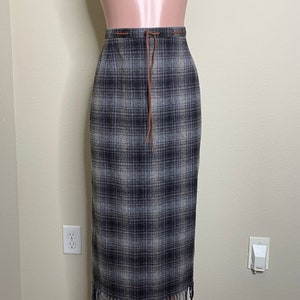 May include: A long, gray and white plaid skirt with a brown leather tie and fringe trim.