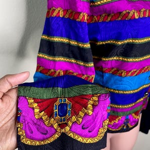 May include: A colorful vintage blazer with a vibrant pattern. The blazer features a mix of blue, purple, and black stripes, accented with gold rope-like trim. The cuff showcases a detailed design with a jewel-like centerpiece, adding a touch of elegance.
