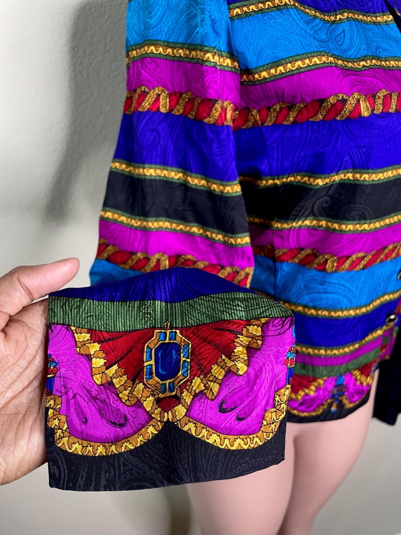 May include: A colorful vintage blazer with a vibrant pattern. The blazer features a mix of blue, purple, and black stripes, accented with gold rope-like trim. The cuff showcases a detailed design with a jewel-like centerpiece, adding a touch of elegance.