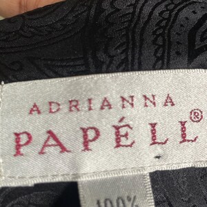 May include: Close-up of a black silk fabric with a paisley pattern. The label reads "ADRIANNA PAPELL" in red letters, with "100% SILK" below. The fabric has a subtle sheen.