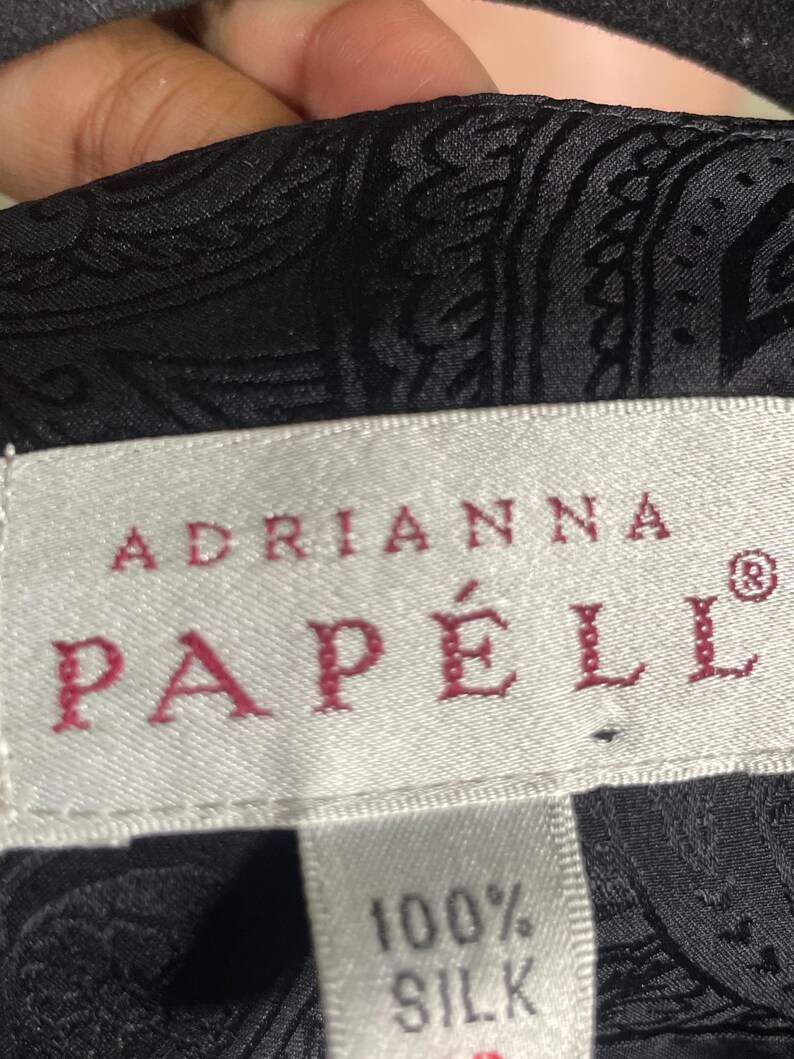 May include: Close-up of a black silk fabric with a paisley pattern. The label reads "ADRIANNA PAPELL" in red letters, with "100% SILK" below. The fabric has a subtle sheen.