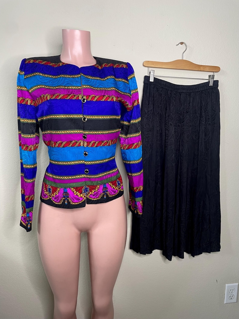 May include: A vintage two-piece suit featuring a colorful, patterned jacket and a black skirt. The jacket has a button-up front and a vibrant design with blue, purple, and red stripes. The skirt is a solid black color.