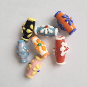 May include: A selection of vibrant glass beads in shades of orange, blue, red, and pink. Each bead is decorated with a floral pattern in contrasting colours. The beads are cylindrical with rounded ends, ideal for jewellery making.