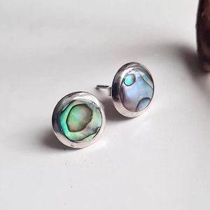 May include: A pair of round stud earrings with iridescent abalone shell inlays. The earrings have a silver-coloured bezel setting. The abalone shell displays shades of green, blue, and purple. The earrings are displayed on a white surface.