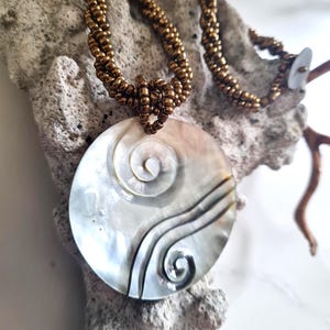 May include: A mother-of-pearl pendant necklace with a large, round pendant. The pendant has a spiral and wave design with dark lines. The necklace has a bronze-coloured beaded chain. The necklace is displayed on a piece of coral.