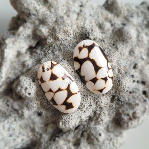 May include: Two oval-shaped pendants with a white base and a dark brown, organic pattern. Each pendant has a small hole at the top for stringing. The pendants are displayed on a textured, gray surface.