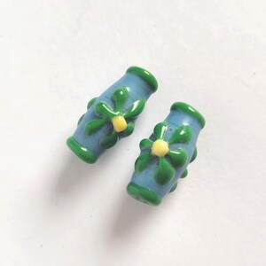 May include: Two blue glass beads with green and yellow floral designs. The beads have a cylindrical shape with green rims and are decorated with green leaves and yellow centers, perfect for jewelry making.