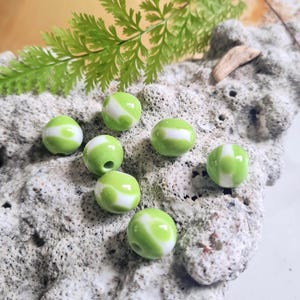 May include: Seven round, green and white beads with a swirled pattern. The beads are a light green colour with white swirls. The beads are on a textured, light grey surface with a fern and a piece of driftwood.