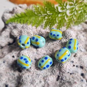 May include: A selection of light blue glass beads, each with yellow stripes and blue polka dots. The oval-shaped beads have a glossy finish. They are displayed on a textured, light grey surface with green foliage in the background.