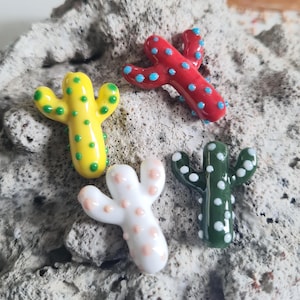 May include: Four colorful glass cactus figurines. One is yellow with green dots, another is red with blue dots, a third is white with pink dots, and the last is green with white dots. The cacti are arranged on a textured, gray surface.