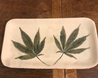 May include: Cream-colored rectangular ceramic tray with two large, detailed green cannabis leaves painted on the surface. The leaves are centered and slightly angled, with visible veins and shading. The tray has rounded corners.