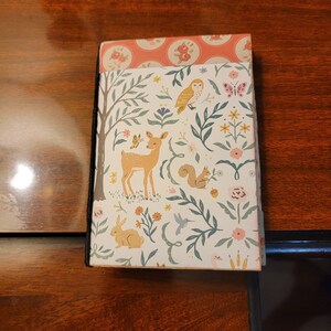 May include: A small journal with a woodland theme. The cover features a deer, squirrel, owl, and rabbit amidst floral and leafy patterns. The top edge has a pink floral design, and the binding is black.