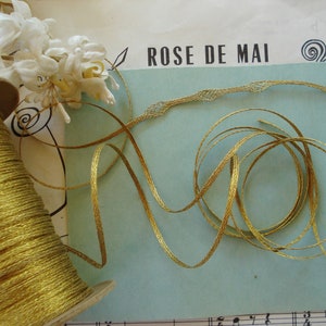 May include: A spool of gold thread with a gold ribbon with a delicate lace pattern. The ribbon is on a blue background with the text "ROSE DE MAI" printed on it.