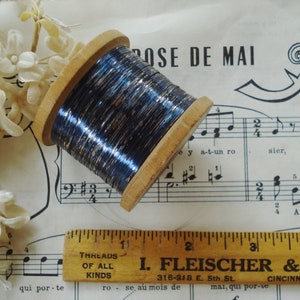 May include: A spool of metallic blue thread with silver flecks, wound on a wooden bobbin. The spool is sitting on a sheet of music with the words "Rose de Mai" printed on it.