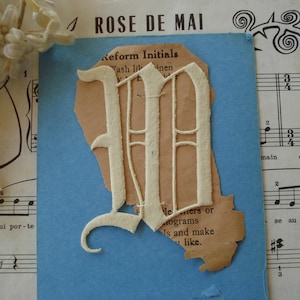 May include: A white felt letter M in a decorative script font, cut out from a piece of brown paper. The letter is on a blue background. The text "Reform Initials" is visible on the paper.