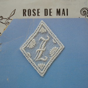 May include: White lace applique in the shape of a diamond with the letter "I" in the center. The lace is delicate and intricate, with a floral design. The applique is perfect for adding a touch of elegance to any project.