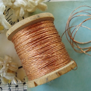 May include: A spool of copper-colored metallic thread wound around a wooden bobbin. The thread is shiny and has a slight shimmer. The spool is sitting on a blue surface.