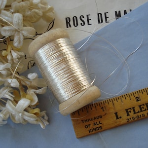 May include: A wooden spool of silver thread is positioned on a light blue surface, alongside a wooden ruler and a cluster of white flowers. The thread is tightly wound. The ruler has black markings and is approximately 7.6 cm long.