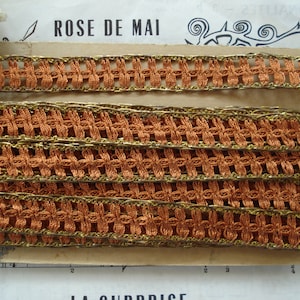 May include: A close-up of a brown and gold trim lace with a woven pattern. The lace is on a piece of cardboard with the text "ROSE DE MAI" printed on it.