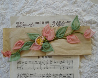 May include: A pink and green silk flower embellishment with the text "ROSE DE MAI" and "LA SURPRISE" on the background.