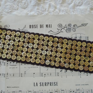 May include: A brown fabric strip with a scalloped edge is covered with a grid of small, round, beige buttons. The fabric strip is lying on a sheet of music with the text "ROSE DE MAI" and "LA SURPRISE" printed on it.
