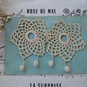 May include: Two off-white, star-shaped crochet doilies featuring a central circular opening and intricate web-like patterns. Each doily has two small, spherical crochet balls hanging from the bottom points by short, thin thread cords.