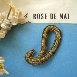 May include: A decorative patch in the shape of the letter J, crafted with metallic gold-toned thread embroidery. The letter features a textured, raised surface with a dense, diagonal stitch pattern. The patch is displayed against a blue background.