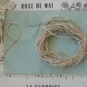 May include: A coiled length of thin, metallic silver cord with a textured, sparkling finish. The cord is arranged in a loose, circular bundle with a single strand extending outward, resting against a light blue rectangular card.