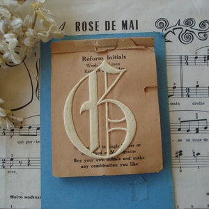 May include: A vintage white paper initial "G" on a beige card with the text "Reform Initials, Wash Linen, Easy to Iron, Sold in single letters or combined as Monograms, Buy your own initials and make any combination you like."