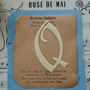 May include: A white felt letter Q with a decorative script design. The letter is on a light brown card with the text "Reform Initials" and instructions on how to use the letters. The card is on a blue background.