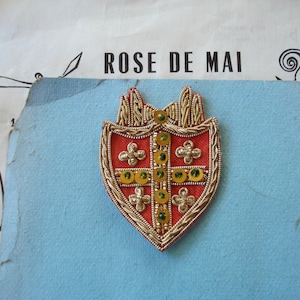 May include: A shield-shaped patch featuring a red fabric background with a gold embroidered cross and border. The design includes four small silver-toned floral emblems and a vertical row of yellow circular accents. The text ROSE DE MAI is visible.